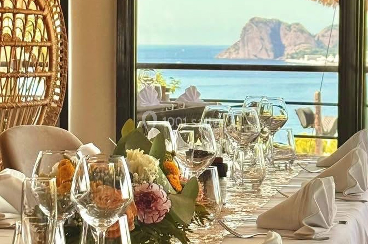 Table dressée avec élégance, ornée de fleurs et de verres, offrant une vue sur la mer et des collines en arrière-plan.