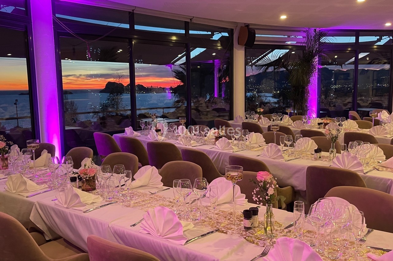 Salle de réception élégamment décorée avec des tables dressées, offrant une vue sur un coucher de soleil en bord de mer.