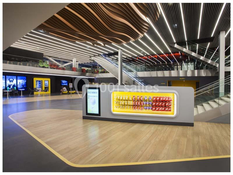 Hall moderne avec sol en bois, plafond ondulé, escalier, et présentoir lumineux de snacks au centre.