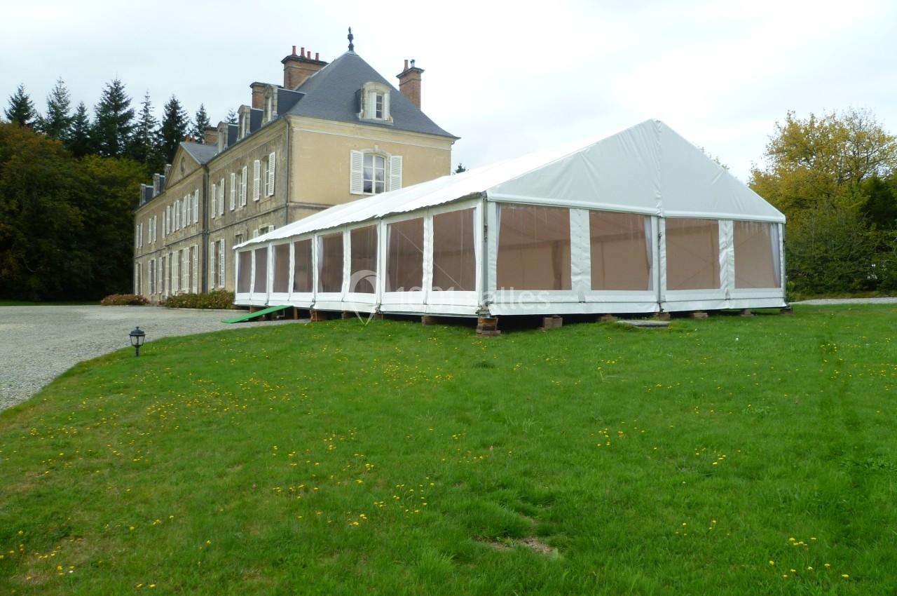 Tente blanche avec parois transparentes installée sur une pelouse devant un grand bâtiment ancien.