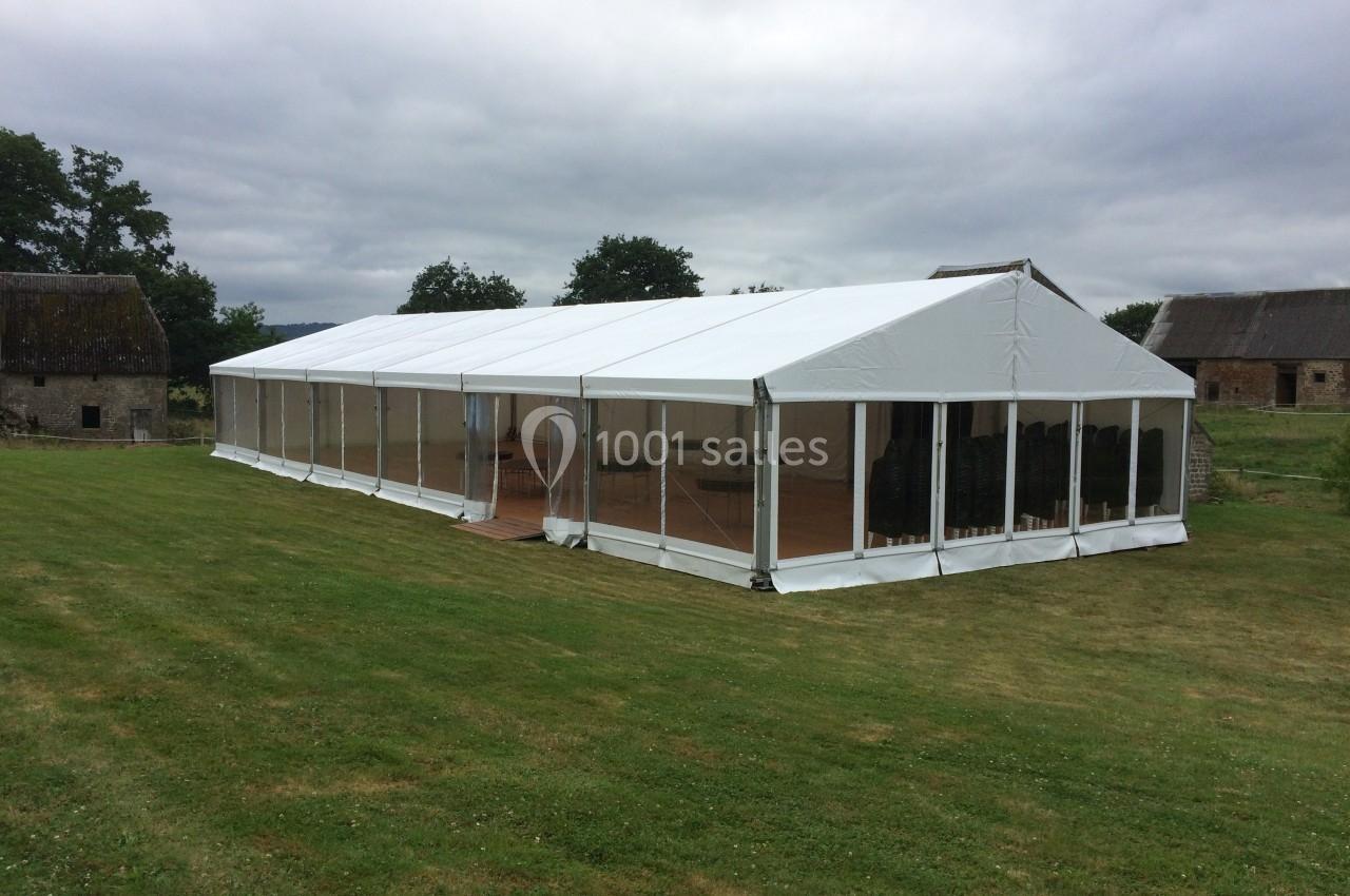 Grande tente blanche avec parois transparentes installée sur une pelouse, entourée de bâtiments ruraux et d'arbres.