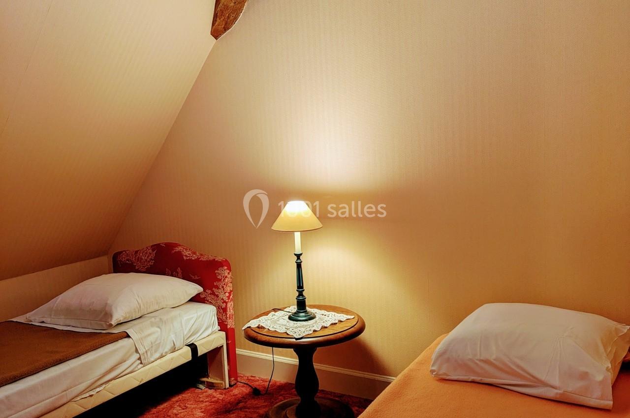 Deux lits simples dans une chambre mansardée avec poutres apparentes, éclairée par une lampe de chevet et un lustre.