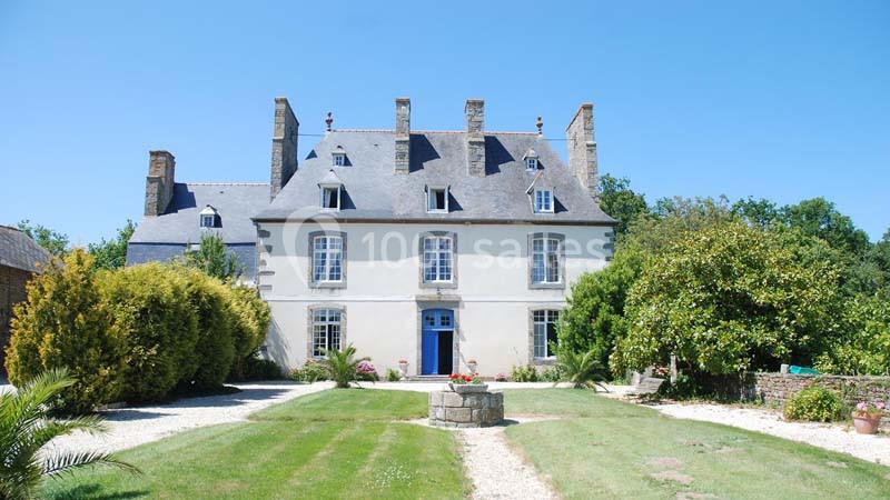 Façade d'un manoir en pierre avec toit en ardoise, entouré de jardins et d'arbres sous un ciel bleu.