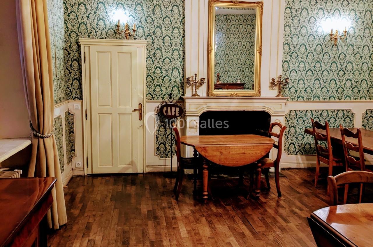 Salle avec parquet en bois, murs ornés de papier peint vert, miroir doré, table ronde et lustre suspendu.