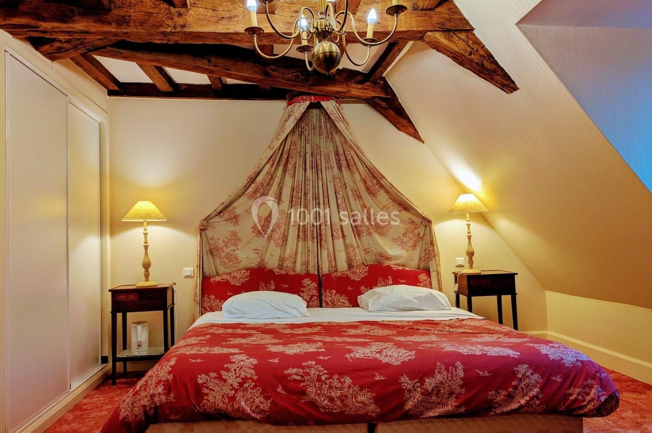 Chambre avec lit double, baldaquin en tissu rouge à motifs, poutres apparentes et éclairage chaleureux.