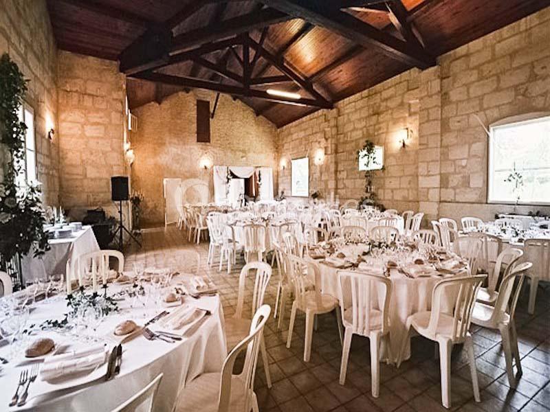 Salle de réception en pierre avec tables rondes dressées, chaises blanches et décoration sobre.