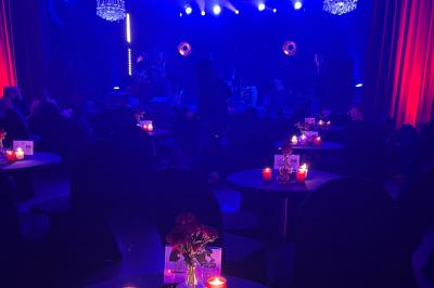 Salle tamisée avec des spectateurs assis à des tables éclairées par des bougies, face à une scène illuminée en rouge et bleu.