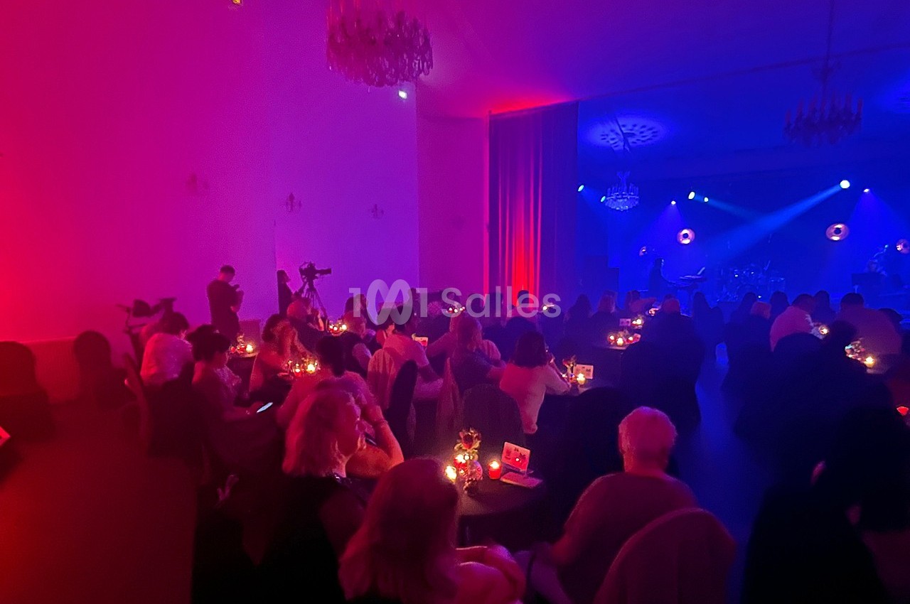 Salle tamisée avec des spectateurs assis à des tables éclairées par des bougies, face à une scène illuminée en rouge et bleu.