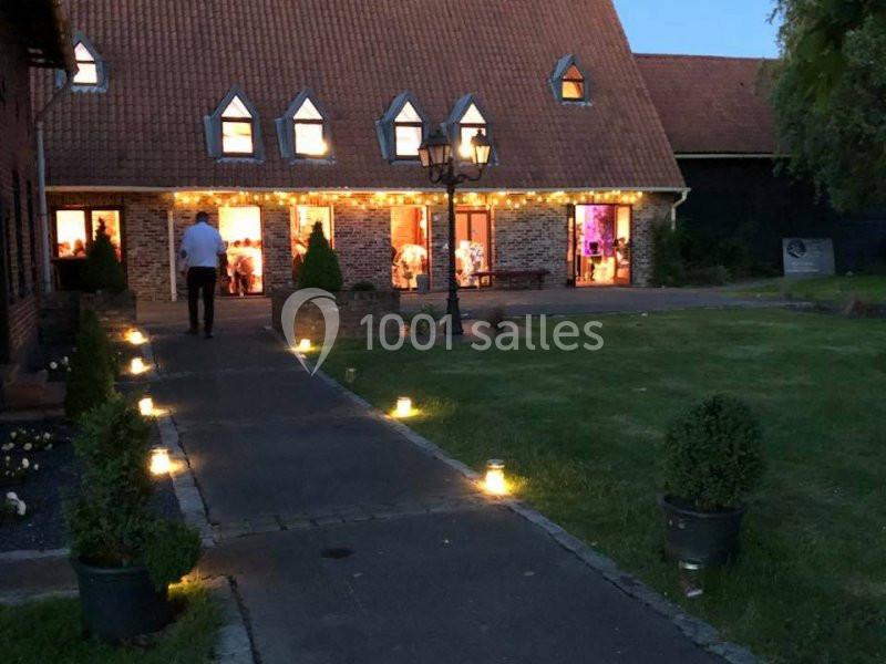 Allée éclairée menant à une maison en briques avec toit en pente, décorée de guirlandes lumineuses en soirée.