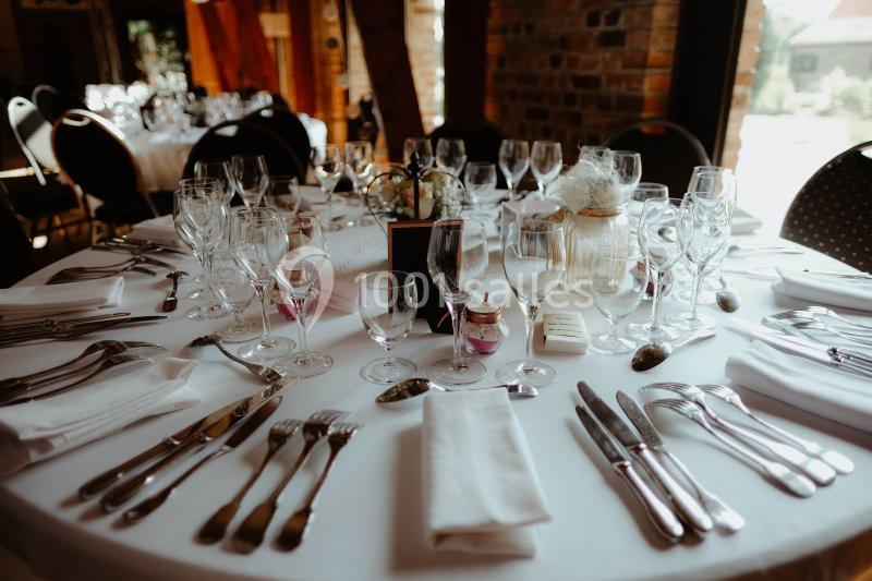 Table ronde élégamment dressée avec nappes blanches, verres, couverts et décoration florale dans une salle lumineuse.