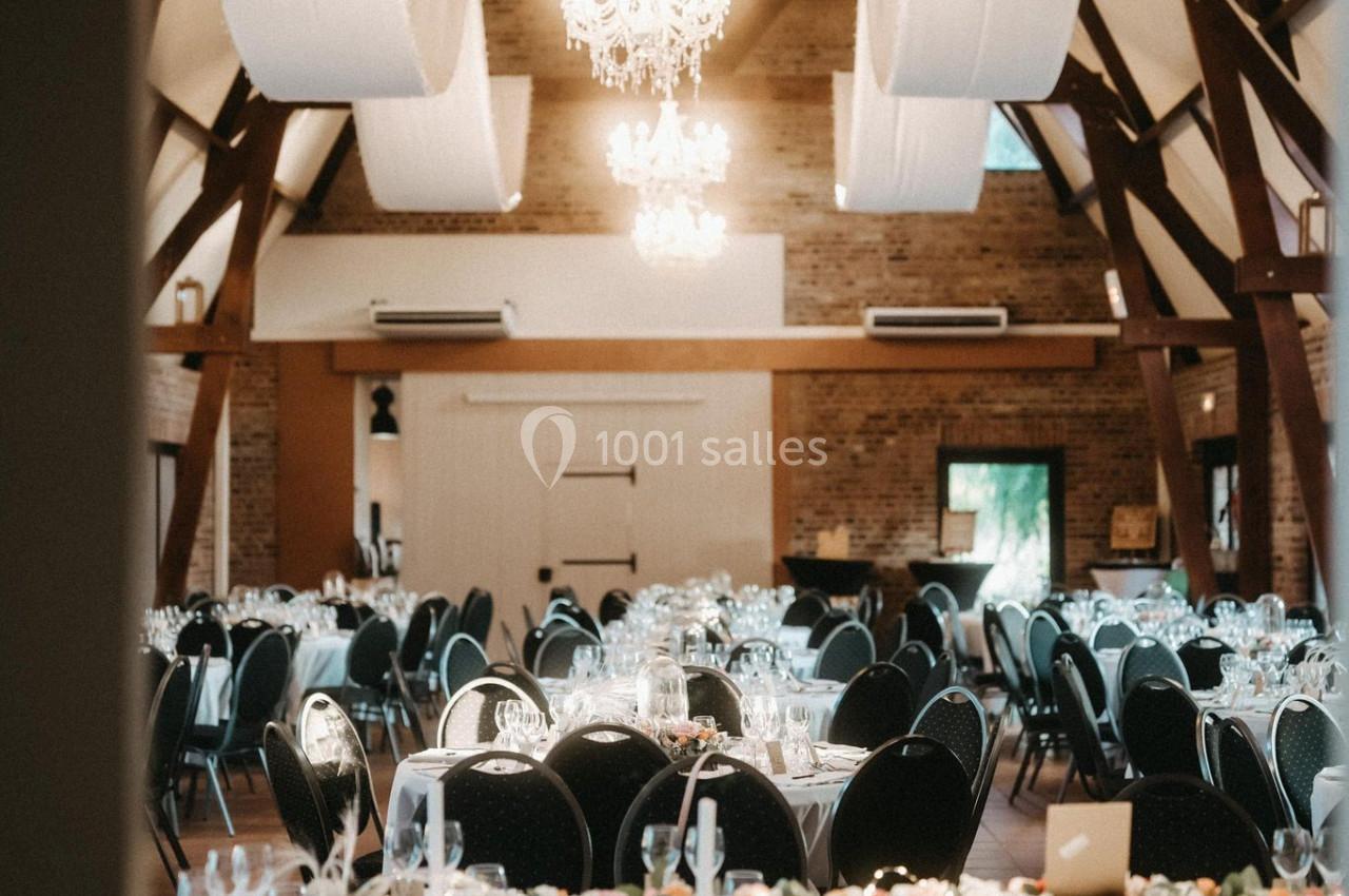 Salle de réception élégante avec tables rondes décorées, chaises noires et lustres suspendus sous une charpente en bois.