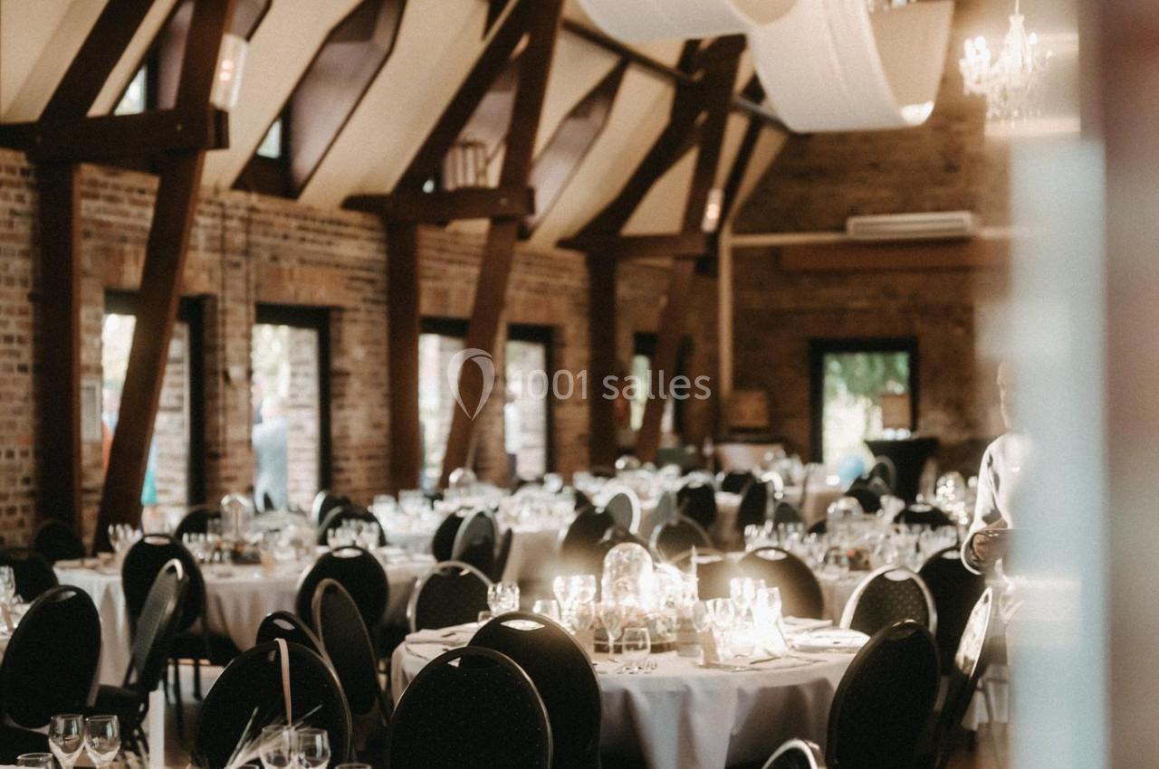 Salle de réception élégante avec tables rondes dressées, nappes blanches, chandeliers et poutres apparentes en bois.