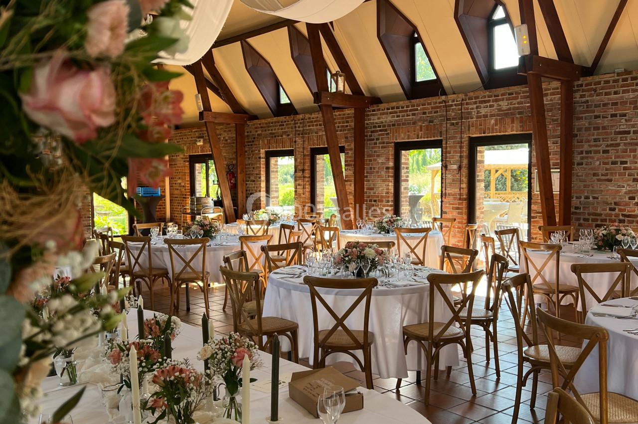 Salle de réception lumineuse avec tables rondes décorées de fleurs, nappes blanches et chaises en bois.