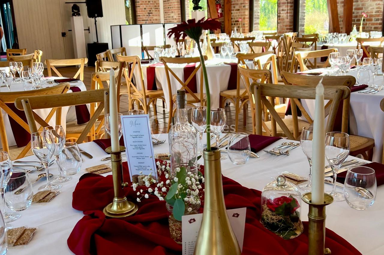 Salle de réception décorée avec des tables dressées, nappes blanches, chemins de table rouges et chandeliers dorés.
