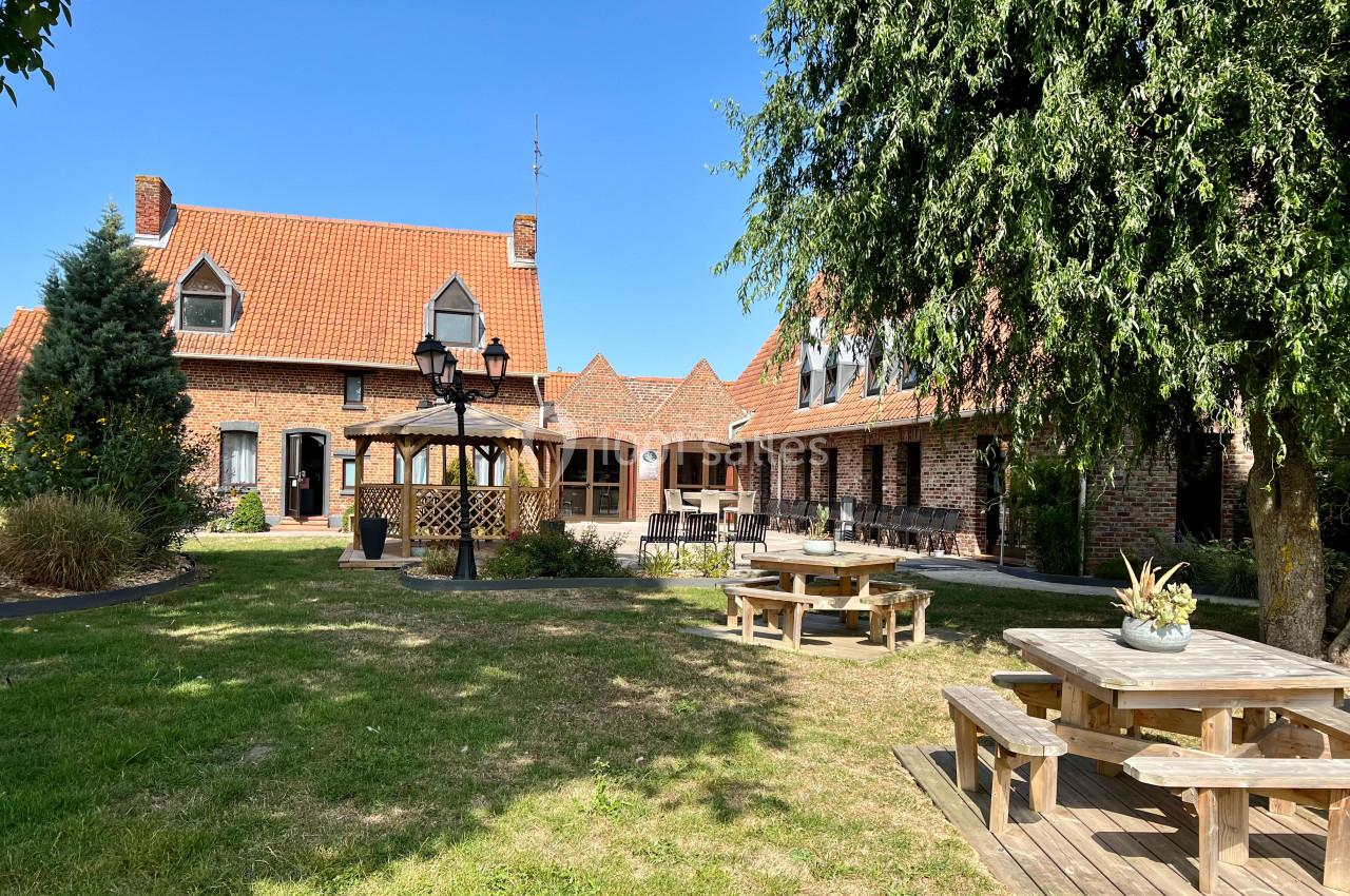 Cour avec jardin, tables en bois, kiosque et bâtiments en briques rouges sous un ciel bleu.