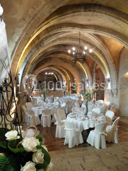 Salle voûtée en pierre aménagée pour un événement, avec tables rondes dressées et décorations florales.