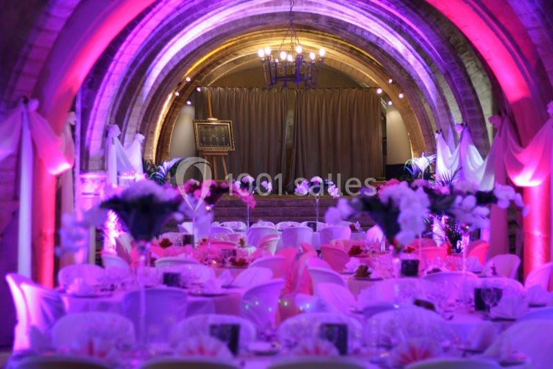Salle voûtée décorée pour un événement, avec tables dressées, fleurs et éclairage rose et violet.