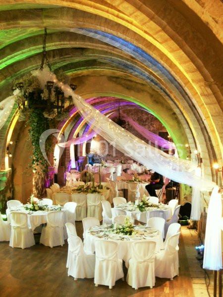 Salle voûtée décorée pour un mariage, avec tables rondes dressées, chaises blanches et éclairage coloré.