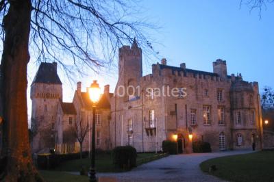 Château en pierre éclairé par des lampadaires au crépuscule, entouré d'arbres et d'une allée gravillonnée.
