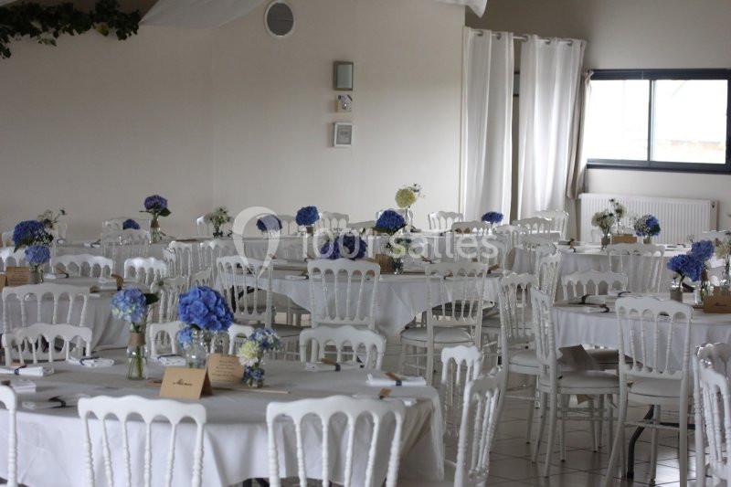 Salle de réception décorée avec des tables rondes, nappes blanches et bouquets de fleurs bleues et blanches.