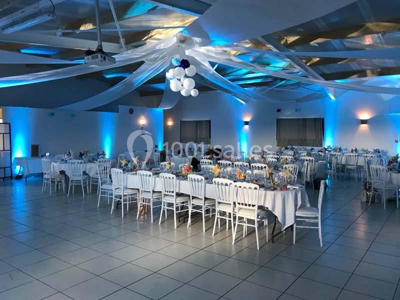 Salle de réception décorée avec des tables dressées, des chaises blanches et un éclairage bleu.