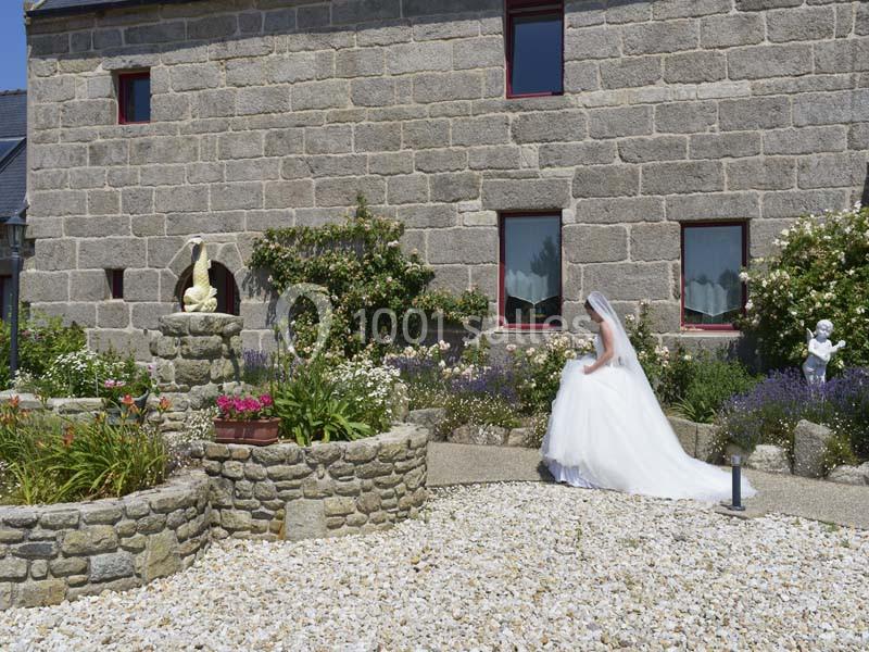 Une mariée en robe blanche marche dans un jardin fleuri devant une maison en pierre avec des statues décoratives.