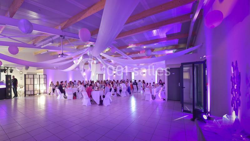 Salle de réception décorée de voilages blancs et éclairée de lumières violettes, avec des invités assis à des tables.