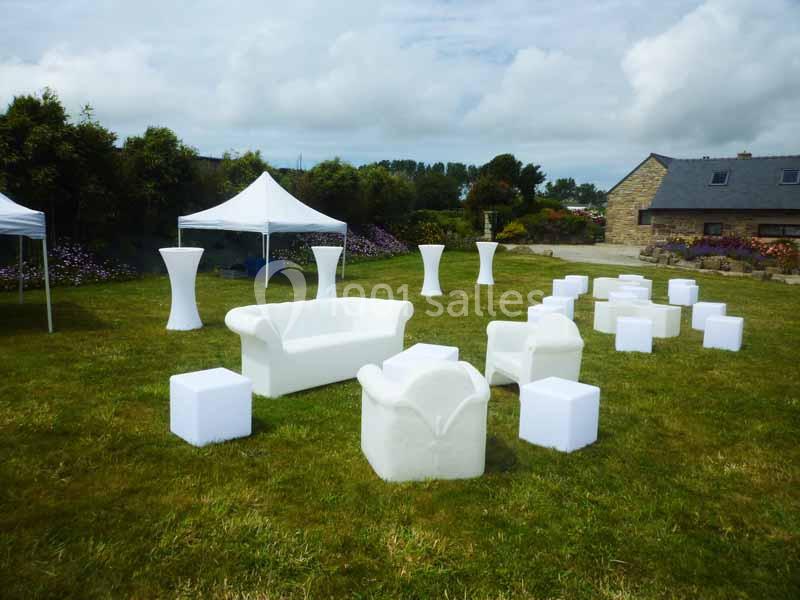 Mobilier blanc moderne disposé sur une pelouse, avec des tentes et un bâtiment en pierre en arrière-plan.