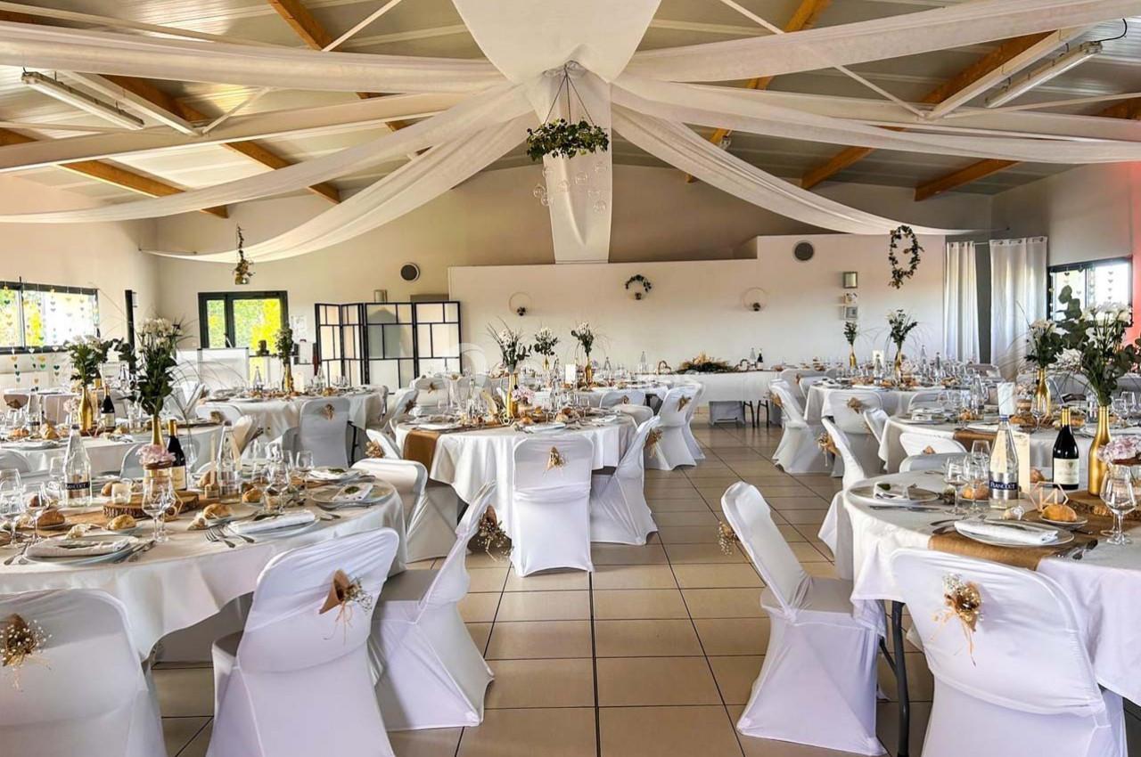 Salle de réception décorée pour un événement, avec tables rondes dressées, nappes blanches et ornements floraux.