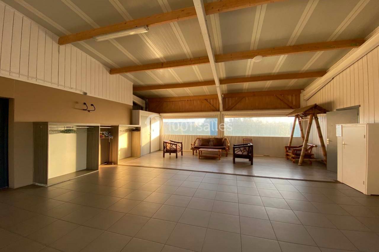 Salle spacieuse avec plafond en poutres apparentes, mobilier en bois, grandes fenêtres et sol carrelé.