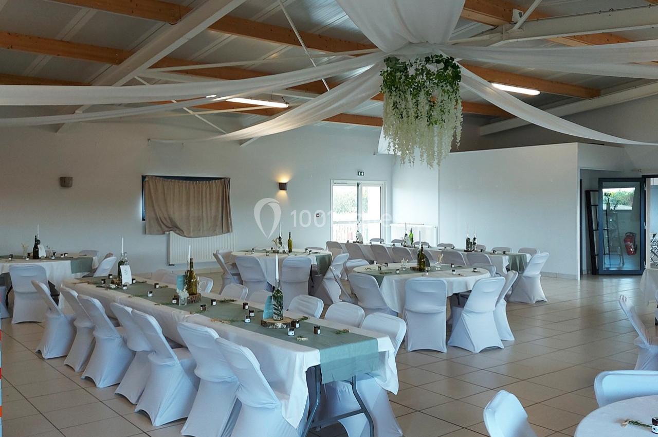 Salle de réception lumineuse avec tables décorées, chaises blanches et plafond orné de drapés et de verdure suspendue.