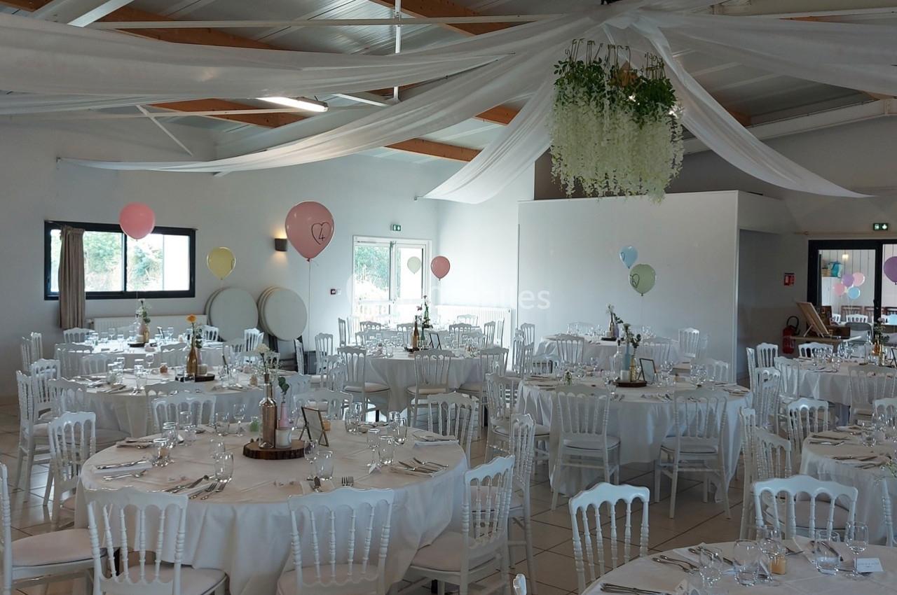 Salle de réception décorée avec des tables rondes, nappes blanches, chaises assorties et ballons colorés suspendus.