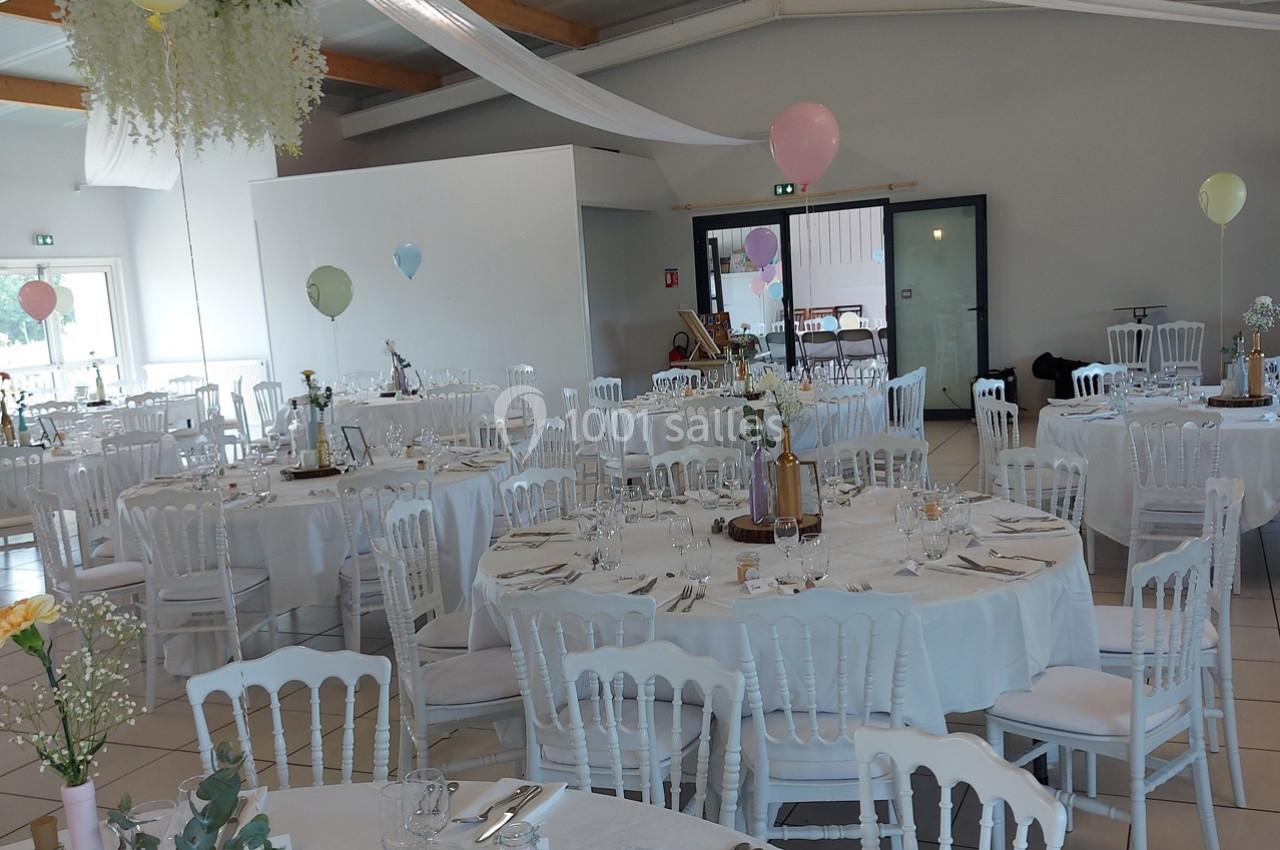 Salle de réception décorée pour un mariage, avec tables rondes dressées, chaises blanches et ballons colorés.
