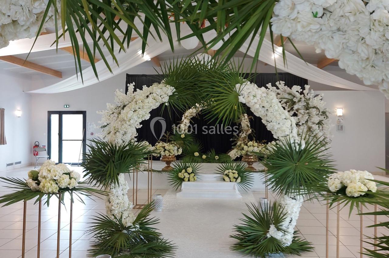Décoration florale blanche et verte avec des arches et des arrangements pour une cérémonie dans une salle lumineuse.