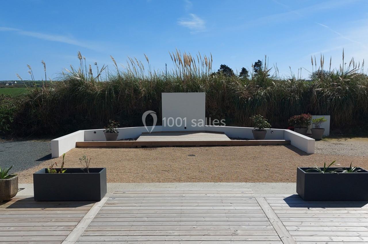 Espace extérieur avec estrade blanche entourée de plantes, bordé par des herbes hautes et un ciel dégagé.