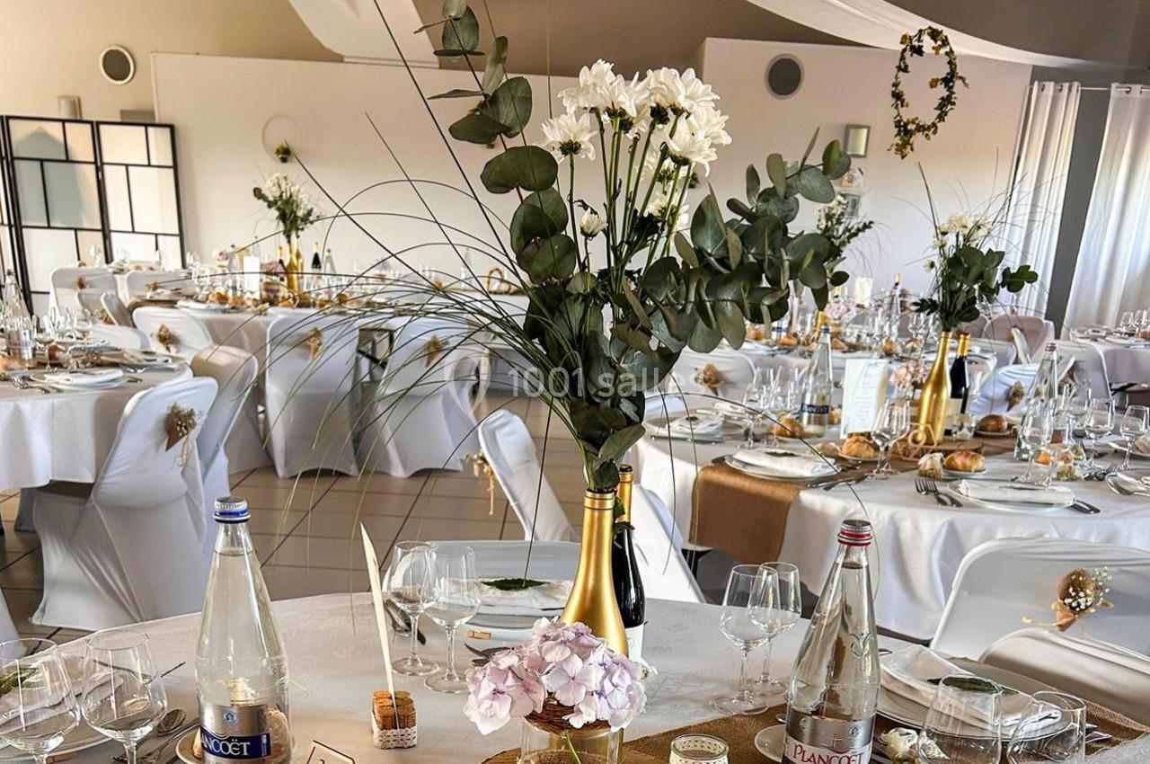 Salle de réception décorée pour un événement, avec tables dressées, fleurs et bouteilles en centre de table.