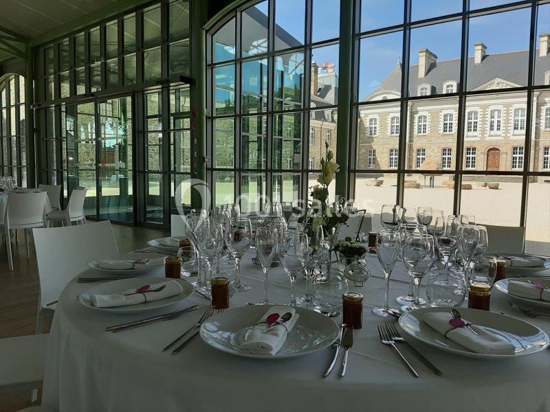Salle lumineuse avec grandes baies vitrées, table ronde dressée avec vaisselle élégante et vue sur un bâtiment en pierre.