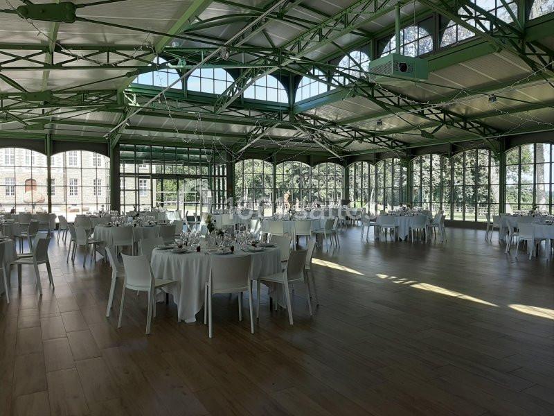 Salle lumineuse avec de grandes baies vitrées, tables rondes dressées et structure métallique verte visible.
