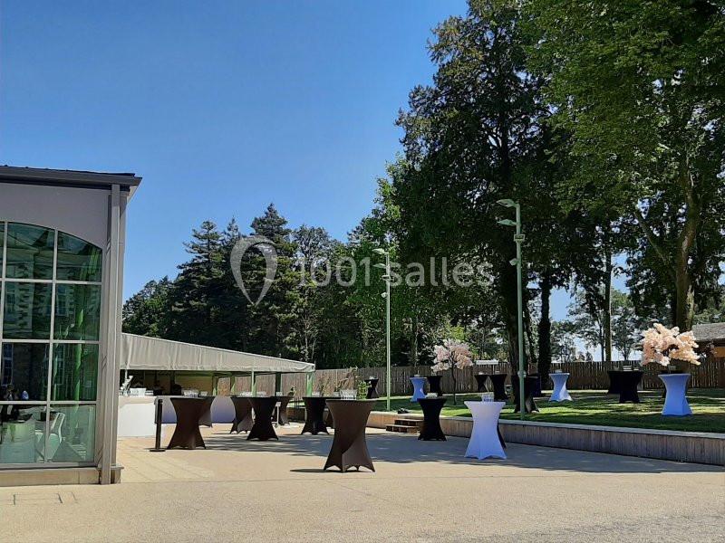Espace extérieur avec tables hautes et décorations florales, entouré d'arbres et d'une structure en verre.