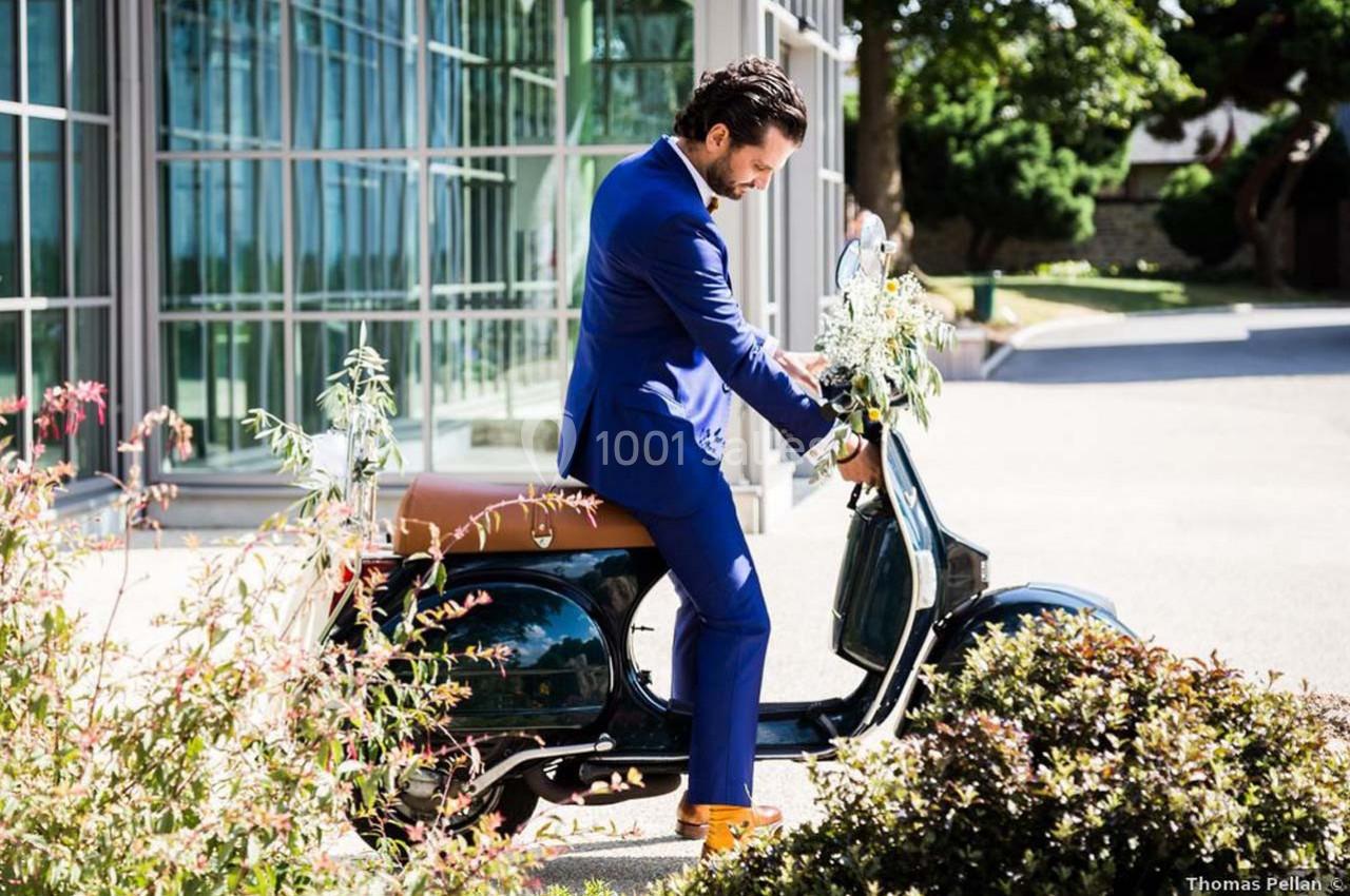 Un homme en costume bleu assis sur un scooter vintage noir, devant un bâtiment vitré et des plantes.