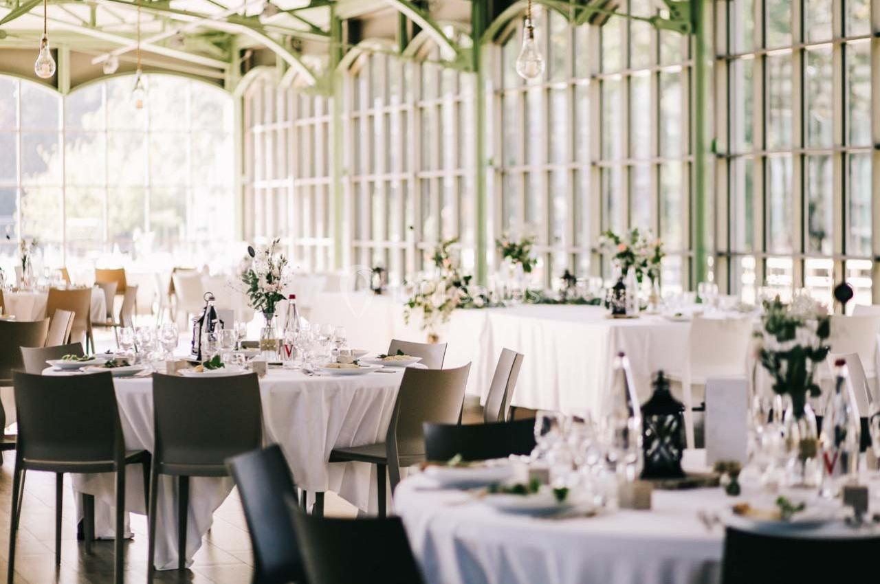 Salle lumineuse avec de grandes baies vitrées, tables rondes dressées pour un événement avec décoration florale.