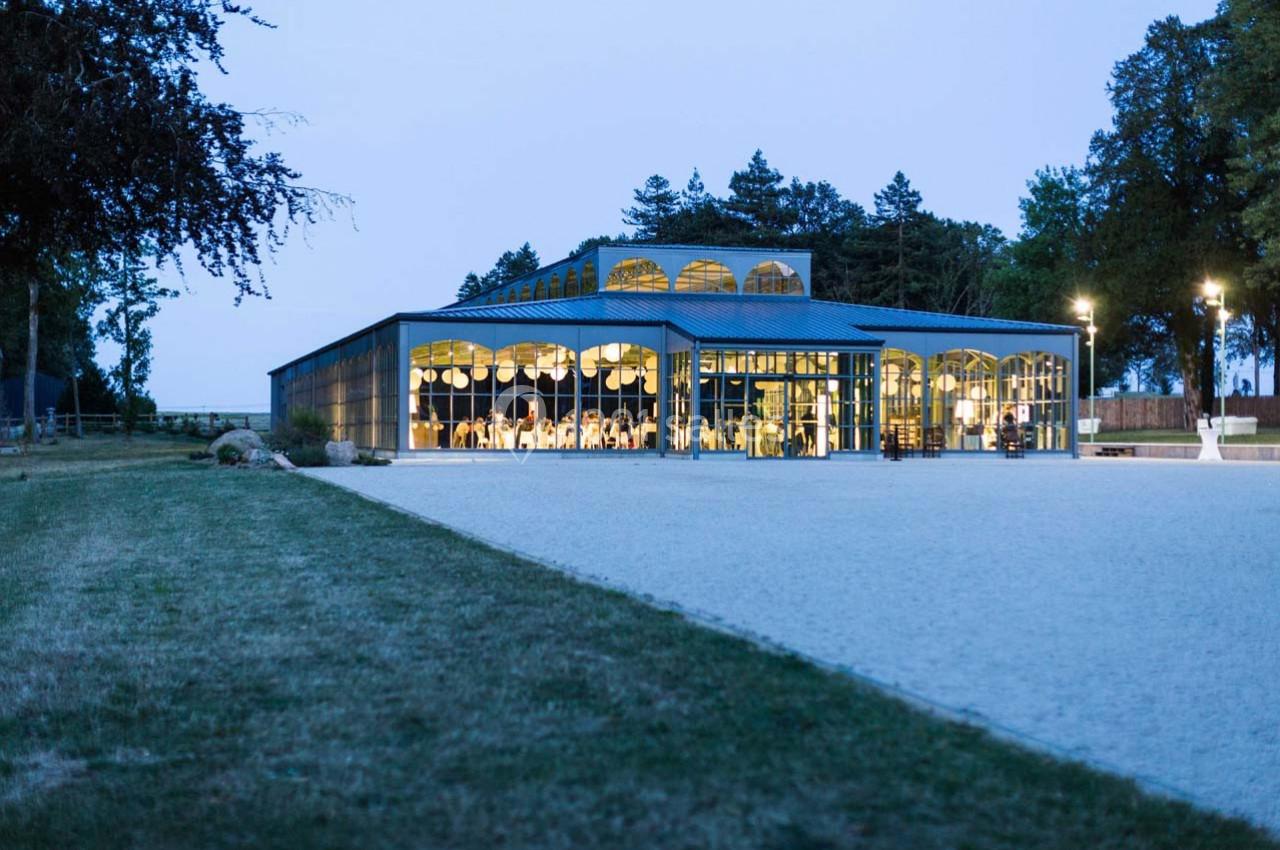 Bâtiment moderne en verre et métal illuminé en soirée, entouré d'arbres et d'une grande cour en gravier.