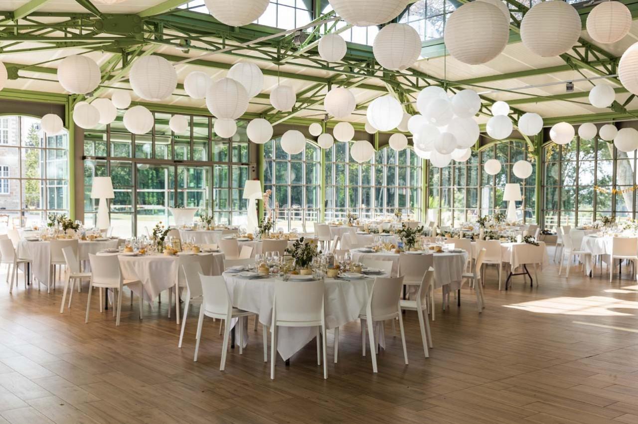 Salle de réception lumineuse avec tables rondes décorées, chaises blanches et lanternes suspendues au plafond.
