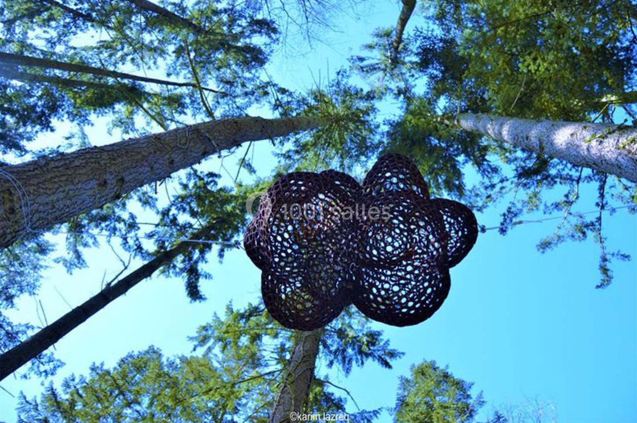 Sculpture suspendue en forme de nuage, composée de mailles sombres, vue depuis le sol entre de hauts arbres.