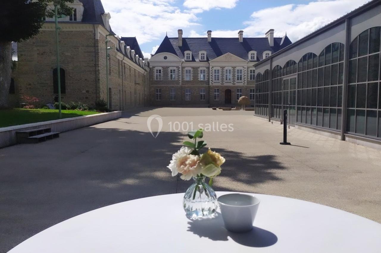 Vase de fleurs et tasse blanche sur une table en extérieur, devant un bâtiment historique sous un ciel dégagé.