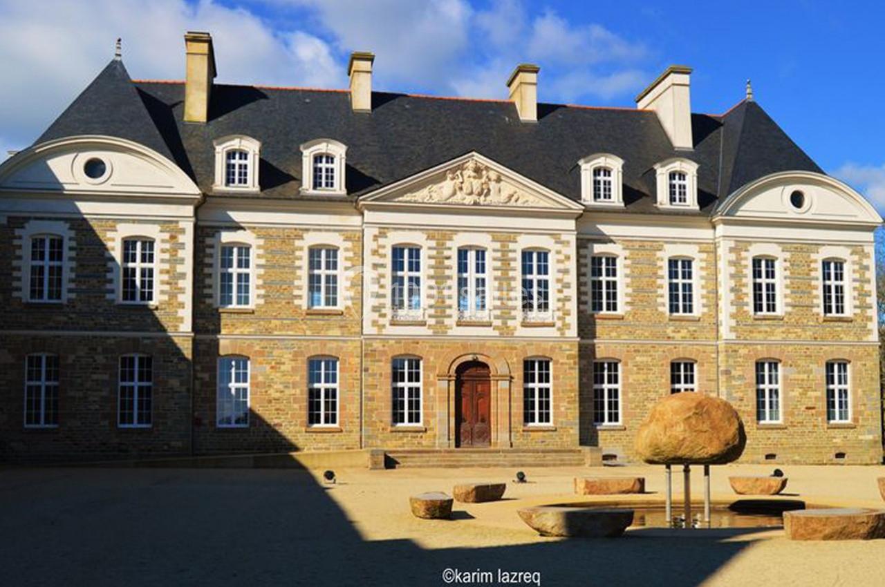 Façade d'un bâtiment historique en pierre avec toit en ardoise, entouré de sculptures en pierre sur une place dégagée.