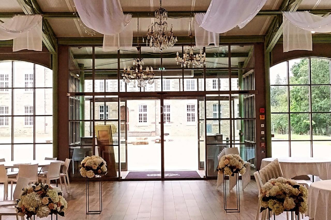 Salle lumineuse avec de grandes baies vitrées, décorée de fleurs blanches et de voilages suspendus.