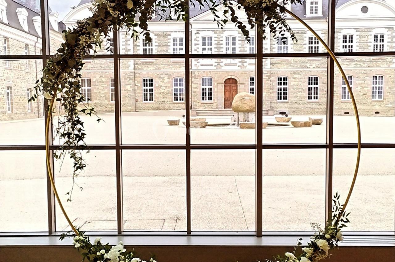 Cercle décoratif fleuri devant une grande fenêtre donnant sur une cour pavée et un bâtiment en pierre.
