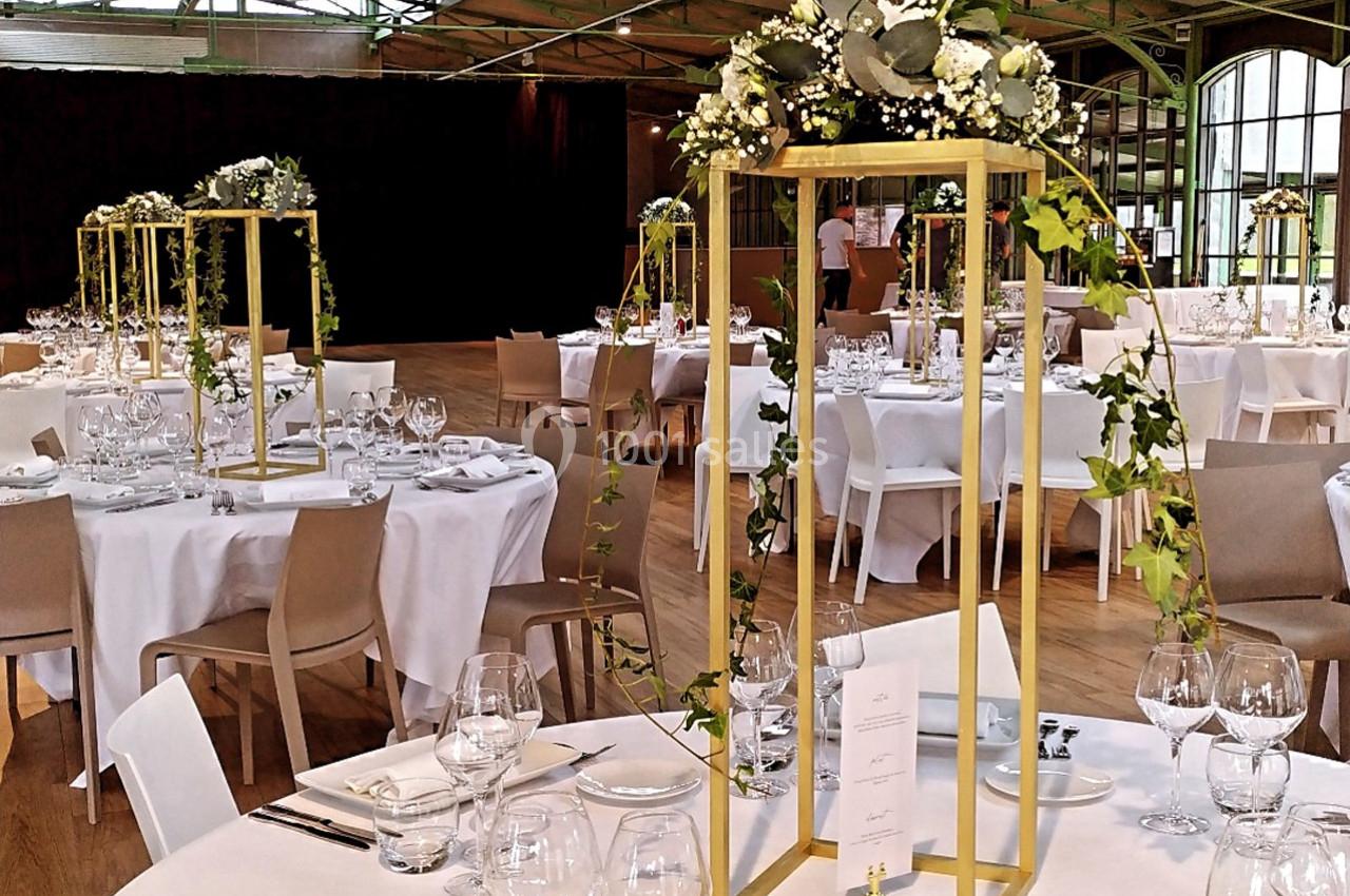 Salle de réception décorée avec des tables rondes, nappes blanches, centres de table dorés ornés de feuillages et fleurs.