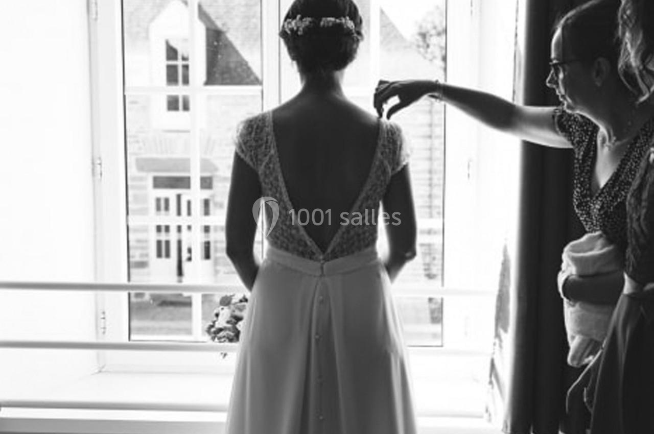 Une mariée de dos en robe blanche près d'une fenêtre, une personne ajuste un détail de sa tenue.